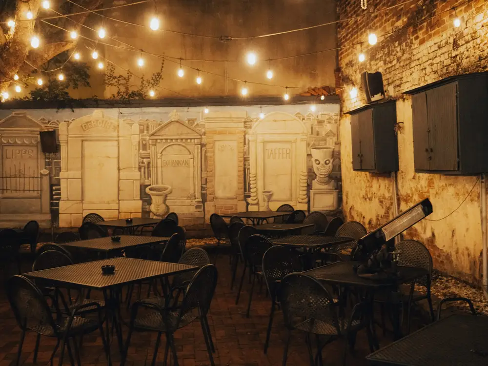 The Backyard at night: iron tables under string lights, in front of a painted wall of old New Orleans tombs, with TVs on the brick wall