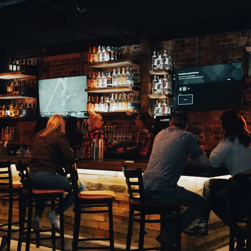 Guests on stools at the long, lit wooden bar, with liquor shelves and TVs behind it
