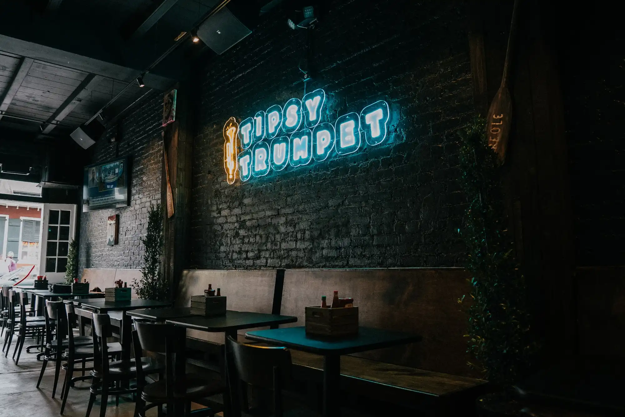 The Tipsy Trumpet dining room: a glowing “Tipsy Trumpet” neon sign on a dark brick wall above booths and tables