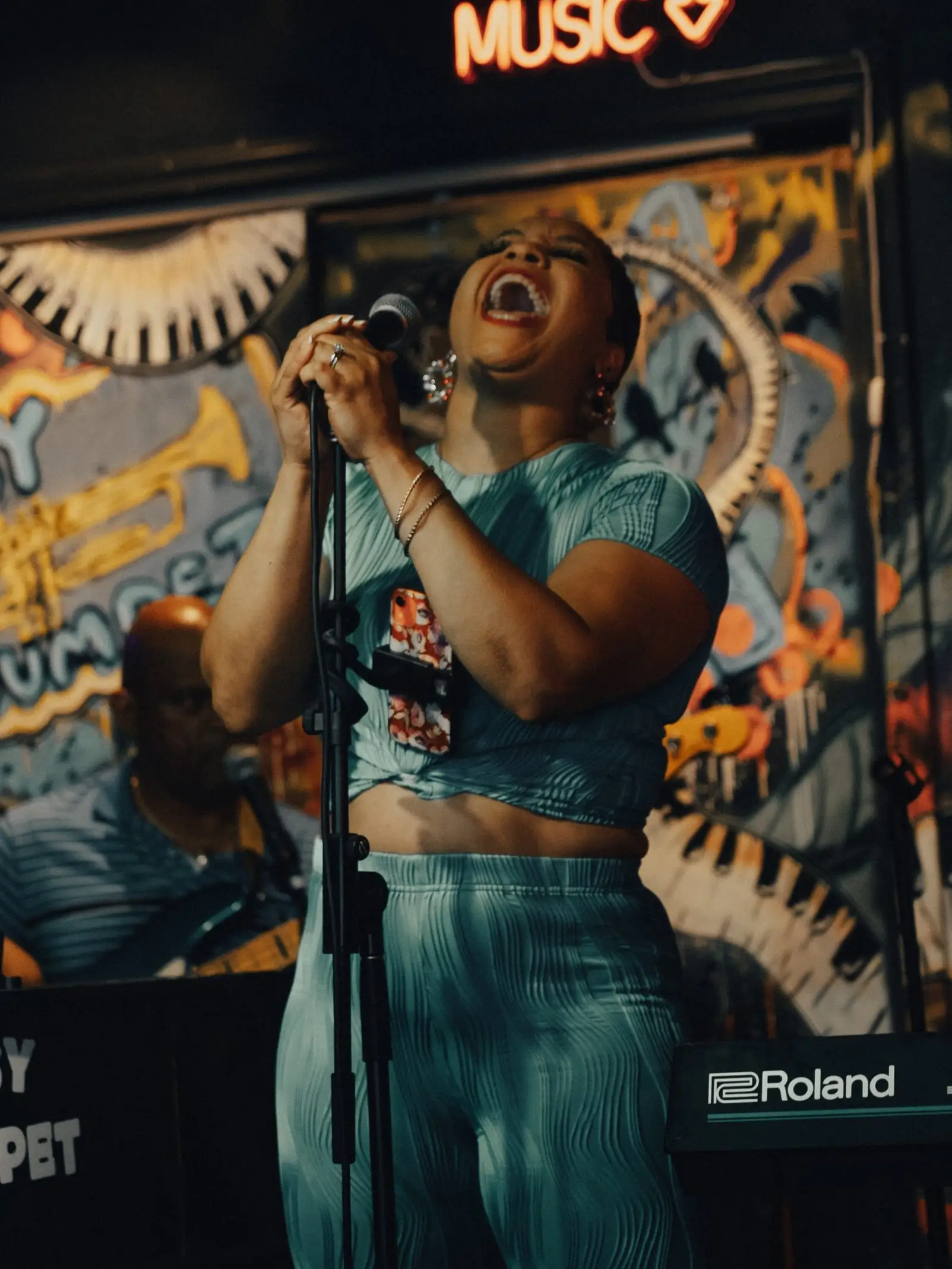 A singer belts out a song under the “LIVE MUSIC” neon sign, in front of the Tipsy Trumpet mural