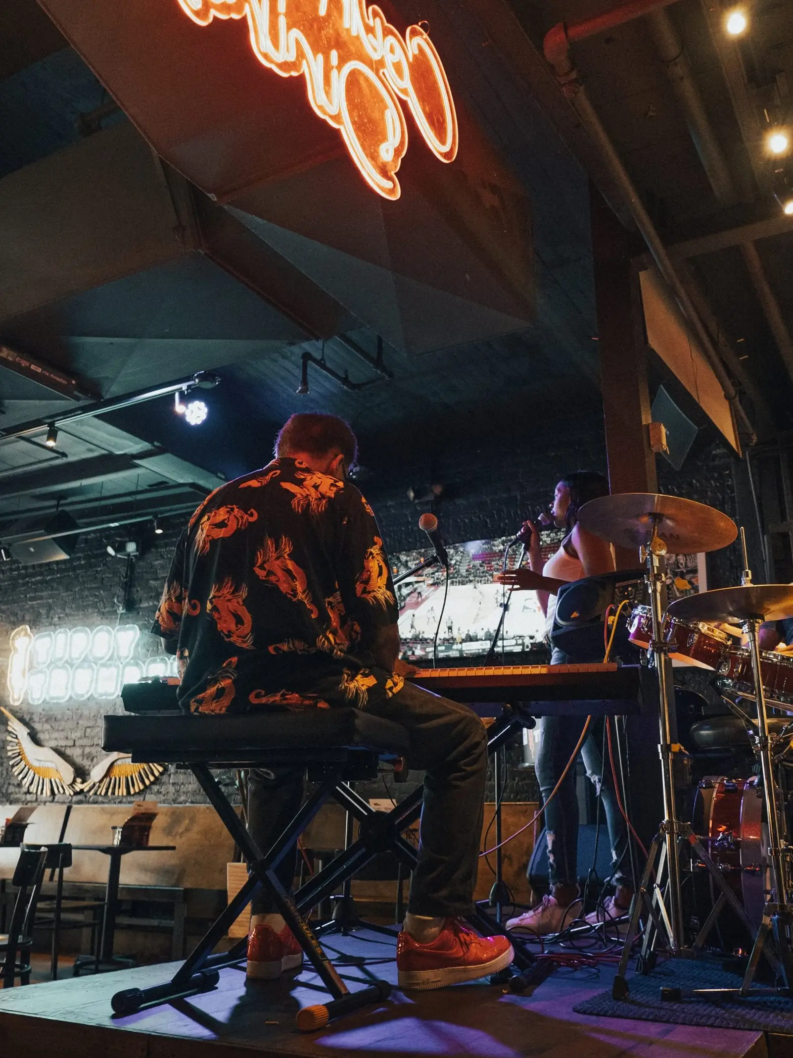 A band on the elevated stage seen from behind the keyboard, with drums and neon lights